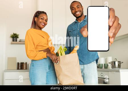 Un couple noir excités déballant un sac de shopping plein d'épicerie, homme montrant un smartphone avec écran vide, maquette Banque D'Images