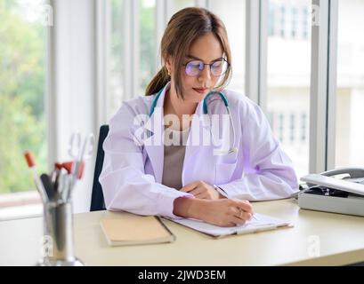 Jeune femme asiatique médecin assis à un bureau et prenant des notes dans le presse-papiers Banque D'Images