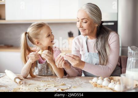 Une vieille grand-mère souriante en tablier enseignant à la petite petite-fille de faire de la pâte, de mouler des biscuits, de préparer de la nourriture Banque D'Images