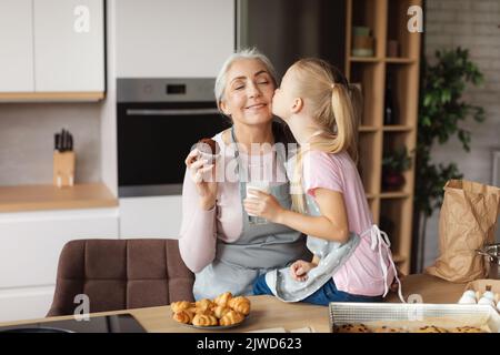 Gaie petite-fille européenne embrasse sa grand-mère sur la joue appréciez les biscuits avec le lait dans la cuisine Banque D'Images