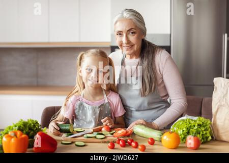 Une petite fille heureuse et une vieille femme en tabliers ont coupé des légumes biologiques pour la salade à l'intérieur minimaliste de la cuisine Banque D'Images