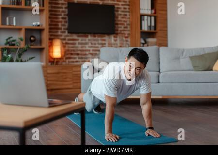 Concept de cours de fitness en ligne. Homme asiatique athlétique planant sur un tapis de yoga à la maison, regardant la vidéo de fitness sur un ordinateur portable Banque D'Images