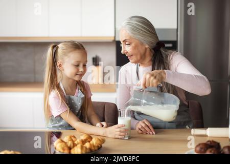 Les bonbons sont prêts. Une bonne grand-mère européenne âgée en tablier verse du lait dans un verre de petite-fille Banque D'Images