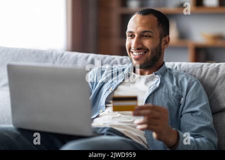 Achats en ligne. Joyeux et charmant Noir homme utilisant un ordinateur portable et une carte de crédit Banque D'Images