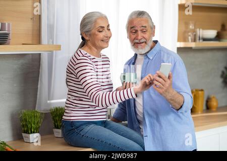 Les conjoints aînés heureux utilisent leur téléphone intelligent ensemble tout en se relaxant dans la cuisine Banque D'Images