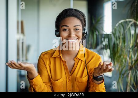 Portrait d'une femme d'affaires noire heureuse dans un micro-casque parlant, faisant des gestes et souriant à l'appareil photo, assis au bureau Banque D'Images