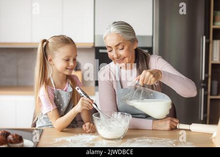 Une bonne grand-mère européenne âgée et une petite petite-fille en tabliers versent du lait dans la pâte Banque D'Images