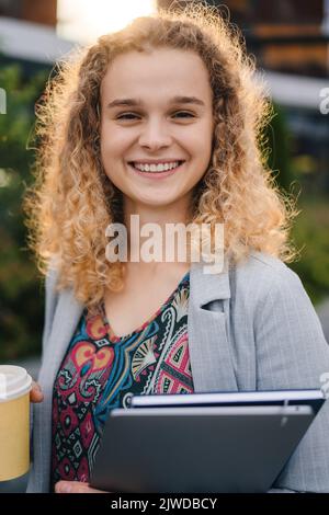 Vue de face d'une femme caucasienne souriante étudiante avec une veste de coiffure en forme de curly, debout à l'extérieur par temps ensoleillé, tenant un café à emporter Banque D'Images