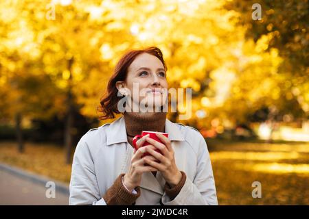 Bonne jeune femme européenne à tête rouge en imperméable avec une tasse de boisson chaude préférée, profitez de la liberté et du temps Banque D'Images