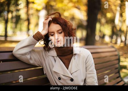 Une jeune femme européenne triste et fatiguée en imperméable est assise sur le banc, pense, souffre de maux de tête et de migraine dans le parc Banque D'Images