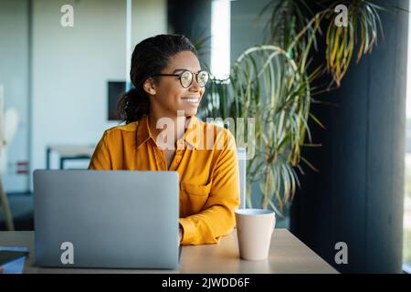 Heureuse femme rêveuse assise sur le lieu de travail avec un ordinateur portable et regardant loin avec le visage de l'expression, l'espace de copie Banque D'Images