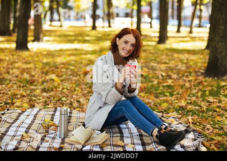 Bonne femme européenne millénaire en imperméable Profitez de votre boisson chaude préférée, vous pourrez vous asseoir sur un plat avec un livre à temps libre Banque D'Images