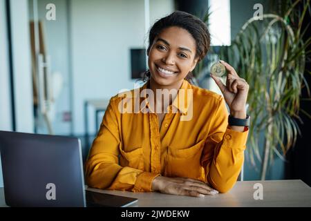 Femme d'affaires noire excitée tenant un bitcoin doré et souriant à l'appareil photo, assise au bureau avec ordinateur portable dans le bureau Banque D'Images
