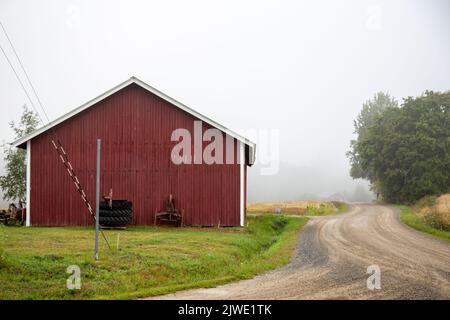 Un grand bâtiment agricole rouge, une traversée tranquille de deux routes de village avec du sable, brouillard en arrière-plan. Banque D'Images