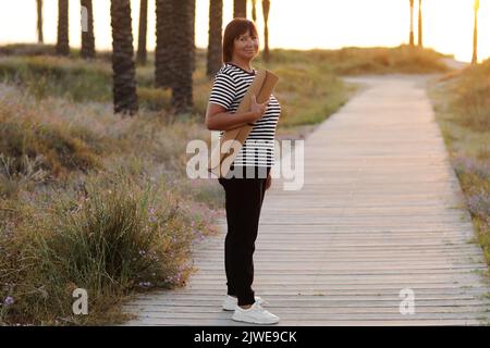 Femme active d'âge moyen avec tapis de yoga et marchant à la plage pour pratiquer le yoga, s'exerçant à l'extérieur avec le sentiment de bonheur au lever du soleil le matin. Co Banque D'Images