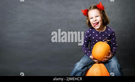 Arrière-plan d'Halloween avec un enfant heureux et de l'espace, enfant drôle tient la citrouille et montre la langue, petite fille mignonne avec des cornes déguissées ayant le plaisir et fait fa Banque D'Images