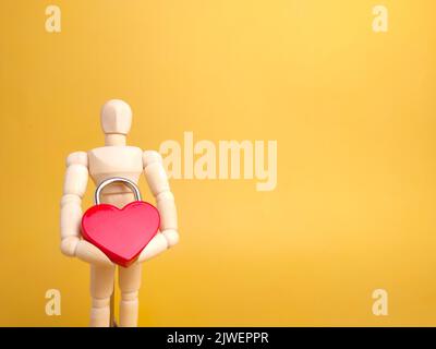 Un mannequin en bois tenant un cadenas rouge en forme d'amour sur fond jaune avec espace pour les copies. Banque D'Images