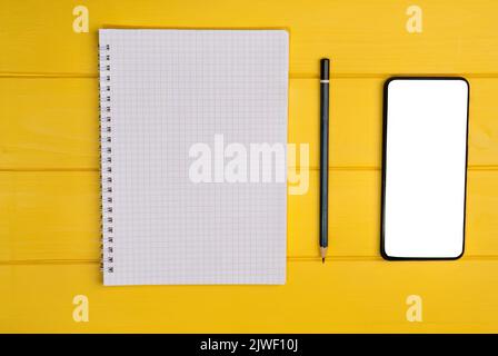 Vue de dessus d'un bloc-notes avec un crayon et un smartphone vierge sur un fond en bois jaune. Banque D'Images