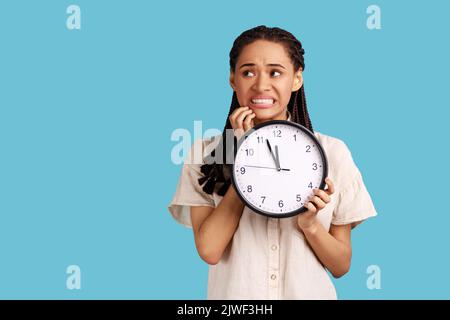 Portrait d'une femme nerveuse impatiente avec des dreadlocks noirs qui mordant ses ongles et tenant une grande horloge murale, date limite, portant une chemise blanche. Studio d'intérieur isolé sur fond bleu. Banque D'Images