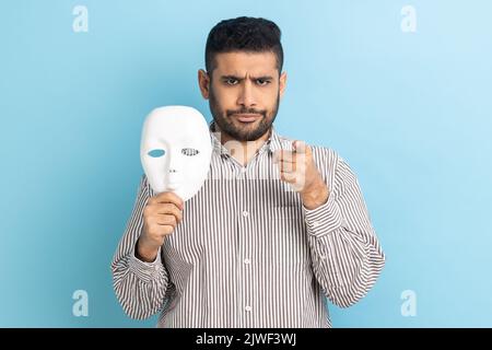 Portrait d'homme d'affaires strict et bossy pointant le doigt vers l'appareil photo et tenant le masque blanc dans sa main, veut changer de personnalité, portant une chemise rayée. Studio d'intérieur isolé sur fond bleu. Banque D'Images