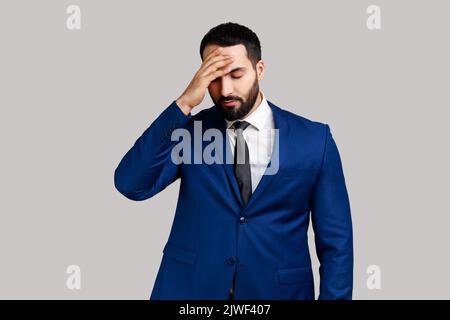 Homme barbu déprimé faisant le geste du visage, se sentir regret et honte au sujet de l'événement oublié, mauvaise mémoire, portant le costume de style officiel. Prise de vue en studio isolée sur fond gris. Banque D'Images