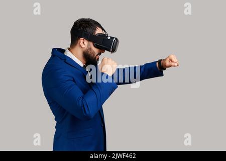 Vue latérale des lunettes de réalité virtuelle MAN sur la tête jouant au jeu de combat, tenant les poings serrés prêts à la boxe, portant un costume de style officiel. Prise de vue en studio isolée sur fond gris. Banque D'Images