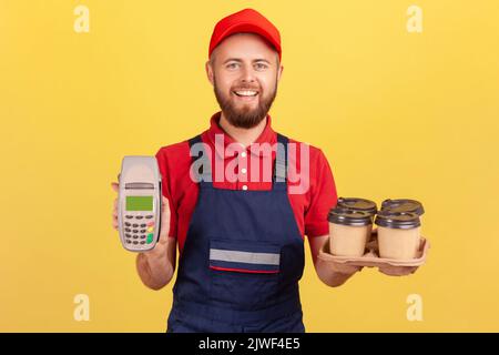 Portrait d'un messager ravie tenant le terminal et le café avec boîte à pizza, regardant l'appareil photo avec une expression heureuse, portant une combinaison et un chapeau. Studio d'intérieur isolé sur fond jaune. Banque D'Images