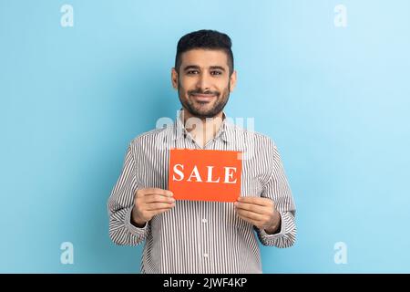 Homme d'affaires souriant tenant la carte avec le mot de vente, se sentant insouciant amusé par des réductions, Black Friday bas prix, portant une chemise rayée. Studio d'intérieur isolé sur fond bleu. Banque D'Images