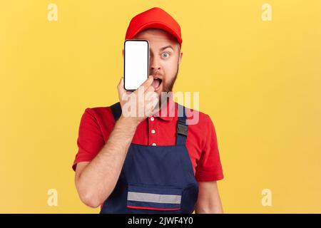 Portrait de l'homme de travail barbu stupéfié en uniforme bleu debout et couvrant l'œil avec téléphone cellulaire avec affichage vide pour le texte promotionnel. Studio d'intérieur isolé sur fond jaune. Banque D'Images