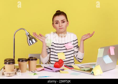 Une femme confuse couverte de notes collantes sur un bureau, avec un ordinateur portable sur un bureau, haussant les épaules, doutant qu'elle ne peut pas prendre de décision. Studio d'intérieur tourné isolé sur fond jaune. Banque D'Images