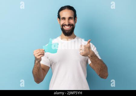 Portrait d'un homme portant un T-shirt blanc blogger montrant les pouces vers le haut le signe de papier bleu, aimer et recommander des postes dans les réseaux sociaux, demandant à évaluer. Studio d'intérieur isolé sur fond bleu. Banque D'Images