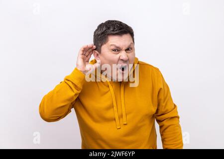 Je ne peux pas vous entendre. Portrait d'un homme charmant et attentif debout avec la main sur l'oreille et qui veut entendre quelque chose, portant un pull à capuche de style urbain. Studio d'intérieur isolé sur fond blanc. Banque D'Images