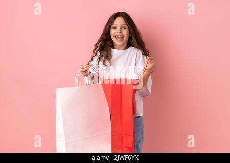 Portrait d'une petite fille excitée et stupéfait portant un T-shirt blanc regardant un appareil photo avec une expression heureuse, tenant des sacs ouverts. Studio d'intérieur isolé sur fond rose. Banque D'Images