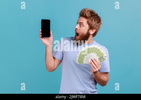 Portrait d'un homme barbu stupéfié tenant des billets en euros et regardant le téléphone portable avec affichage vide, maquette pour la publicité. Studio d'intérieur isolé sur fond bleu. Banque D'Images