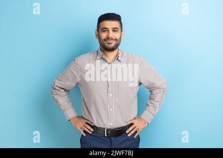 Portrait d'un homme d'affaires à barbe heureux et attrayant avec le sourire tenant les mains sur les hanches, regardant l'appareil photo avec l'expression confiante, portant une chemise rayée. Studio d'intérieur isolé sur fond bleu. Banque D'Images