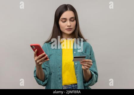 Portrait d'une femme concentrée aux cheveux sombres tenant un smartphone et une carte de crédit, saisissant des données, magasiner en ligne, portant une veste de style décontracté. Prise de vue en studio isolée sur fond gris. Banque D'Images