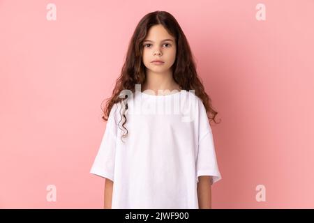 Portrait d'une petite fille sérieuse stricte portant un T-shirt blanc debout regardant l'appareil photo avec l'expression bossy, étant de mauvaise humeur. Studio d'intérieur isolé sur fond rose. Banque D'Images