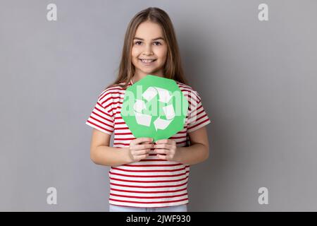 Portrait de charmante petite fille responsable portant un T-shirt rayé tenant dans les mains vert recyclage chanter, écologie concept, regardant l'appareil photo. Prise de vue en studio isolée sur fond gris. Banque D'Images