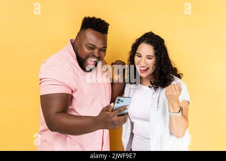 Portrait d'un jeune couple très heureux et excité en vêtements décontractés debout ensemble et regardant le smartphone, les poings serrés, célébrant la victoire. Studio d'intérieur isolé sur fond jaune Banque D'Images