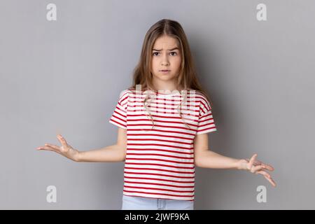 Que voulez-vous ? Portrait d'une petite fille portant un T-shirt rayé debout avec des mains levées et surpris d'expression indignée, demandant quelle raison. Prise de vue en studio isolée sur fond gris. Banque D'Images