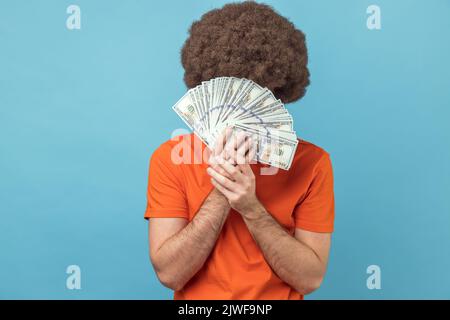 Portrait d'un homme avec une coiffure afro portant un T-shirt orange cache face derrière un bouquet de billets en dollars, personne anonyme tenant de l'argent. Studio d'intérieur isolé sur fond bleu. Banque D'Images
