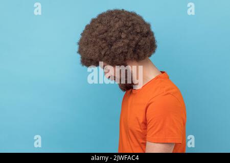 Vue latérale d'un homme mécontent avec une coiffure afro portant un T-shirt orange regardant un appareil photo avec une triste expression faciale, ayant des problèmes. Studio d'intérieur isolé sur fond bleu. Banque D'Images