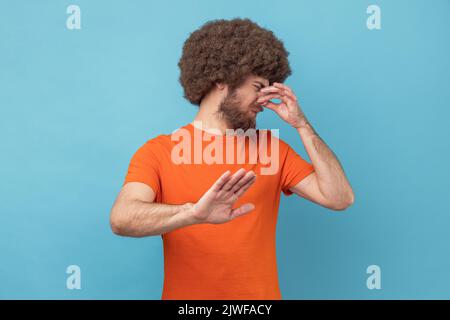 Portrait d'un homme avec une coiffure afro portant un T-shirt orange se détournant et nez dans le dégoût, en évitant l'odeur désagréable de farting, stop geste. Studio d'intérieur isolé sur fond bleu. Banque D'Images