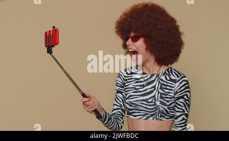 Réaction de Wow. J'ai impressionné la jeune femme avec une perruque brune blogger prenant selfie sur téléphone mobile selfie stick, communiquant appel vidéo en ligne. Adulte élégante fille sur fond beige studio intérieur Banque D'Images