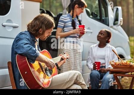 Jeune homme assis sur une chaise et jouant de la guitare pendant le camping dans la nature avec deux filles parlant en arrière-plan Banque D'Images