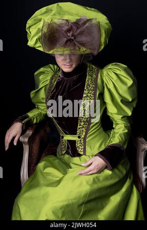 Femme victorienne portant un ensemble de soie verte et assise dans une chaise en velours rouge sur fond noir Banque D'Images