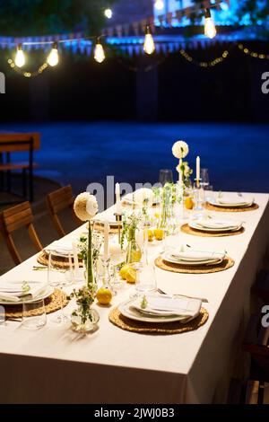 Table longue couverte de nappe blanche avec fleurs de dahlia, bougies, décorations et verres de vin entourés de deux rangées d'assiettes Banque D'Images