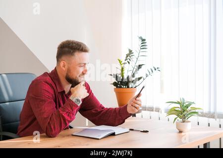 Un journaliste indépendant rouge et décontracté qui a réussi à discuter de la communication vidéo par téléphone tout en étant assis à son bureau et en prenant des notes en riant avec rire. Vidéoconférence avec les partenaires professionnels Banque D'Images