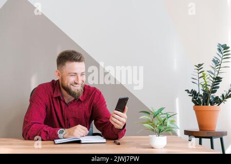 Un journaliste indépendant rouge et décontracté qui a réussi à discuter de la communication vidéo par téléphone tout en étant assis à son bureau et en prenant des notes en riant avec rire. Vidéoconférence avec les partenaires professionnels Banque D'Images