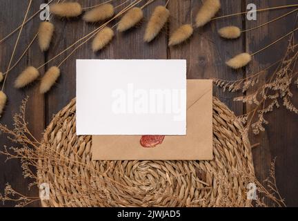 Carte vierge et enveloppe sur le platemat à wattLED sur le bois près de la vue de dessus de l'herbe de queue de Hare. Scène boho avec carte papier horizontale. Bohemian ou proche de natur Banque D'Images